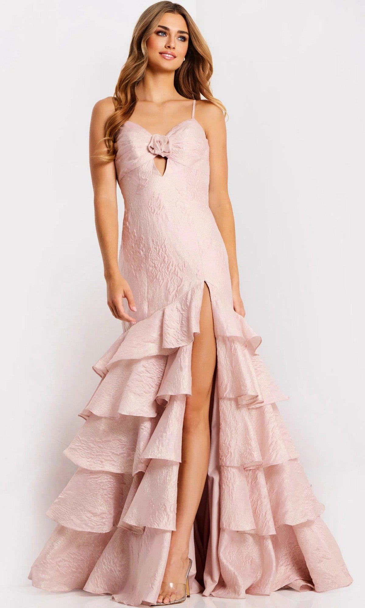 A woman stands against a plain background wearing the Jovani 47329 long prom dress featuring light pink textured fabric, spaghetti straps, a high slit, tiered ruffle skirt, and a small front bow detail.